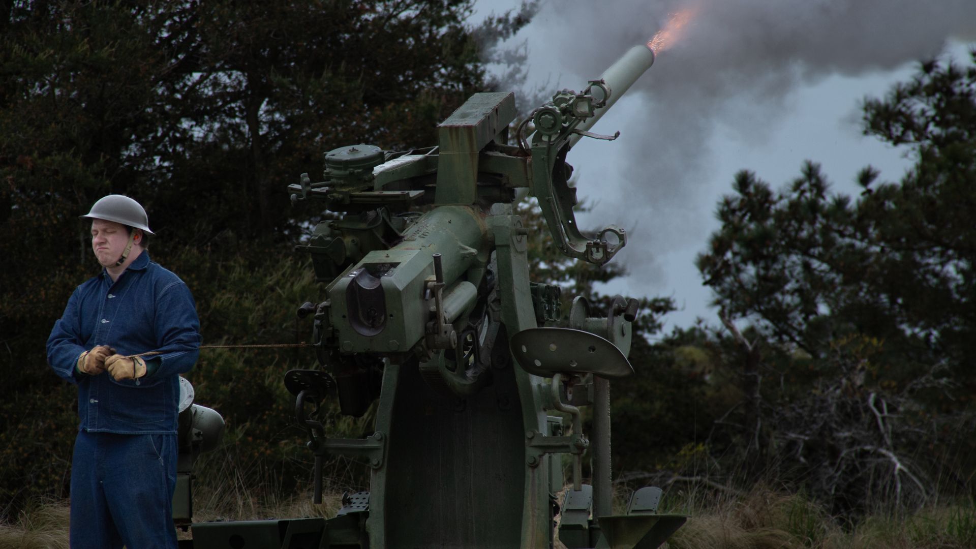 Man in blue coveralls and a white helmet stands beside a large artillery gun as it fires, with smoke billowing in the background.