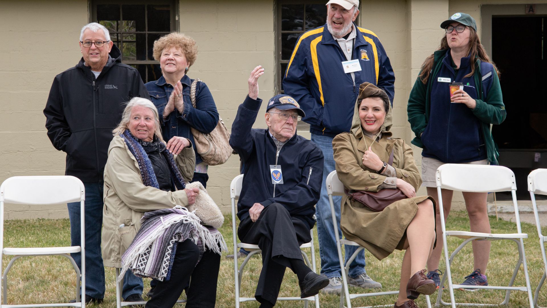 Group of smiling adults seated and standing outside a beige building, watching an event together.