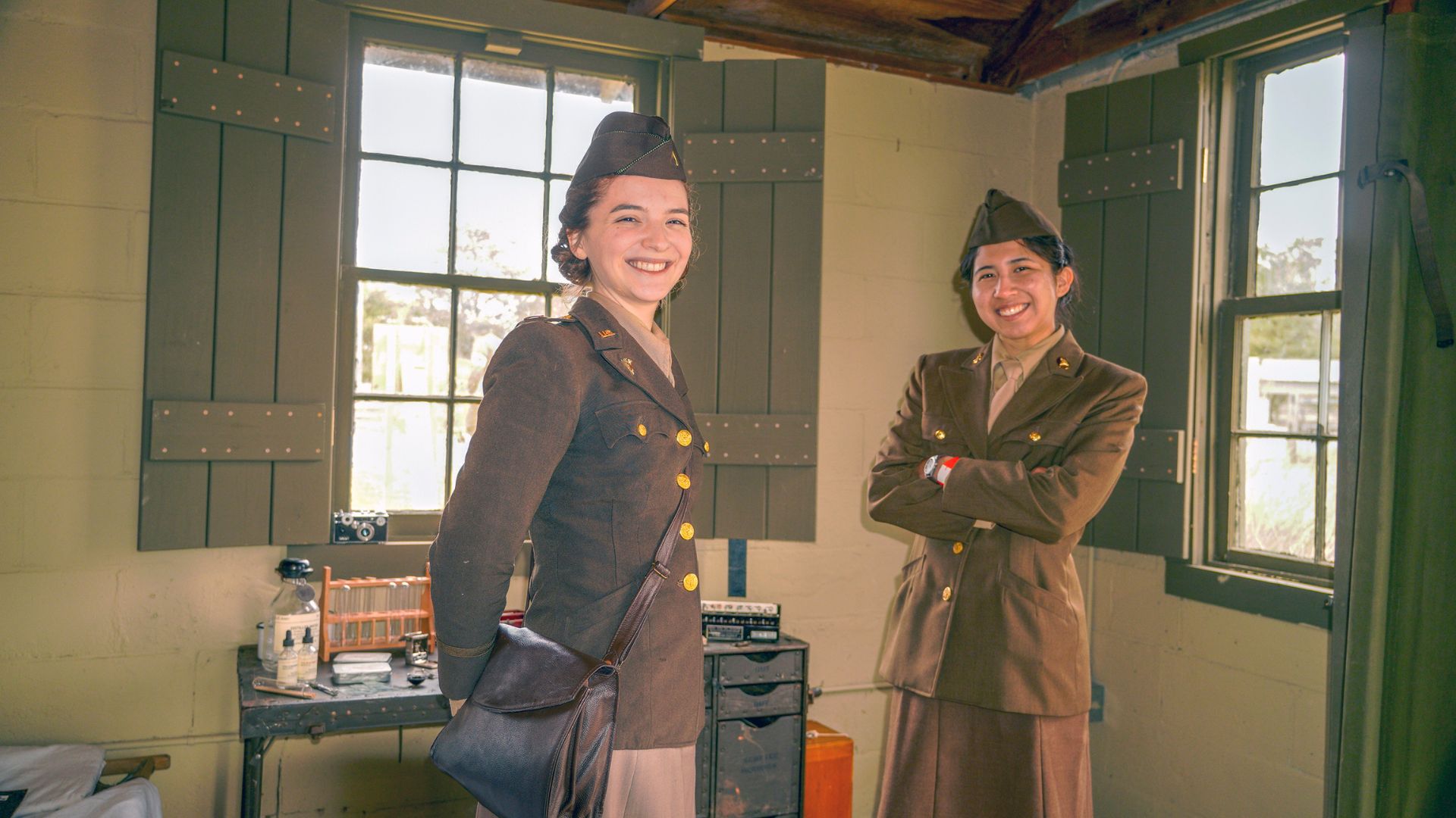 Two women in vintage military uniforms smile and pose in a sunlit, period-style room with green shutters.",