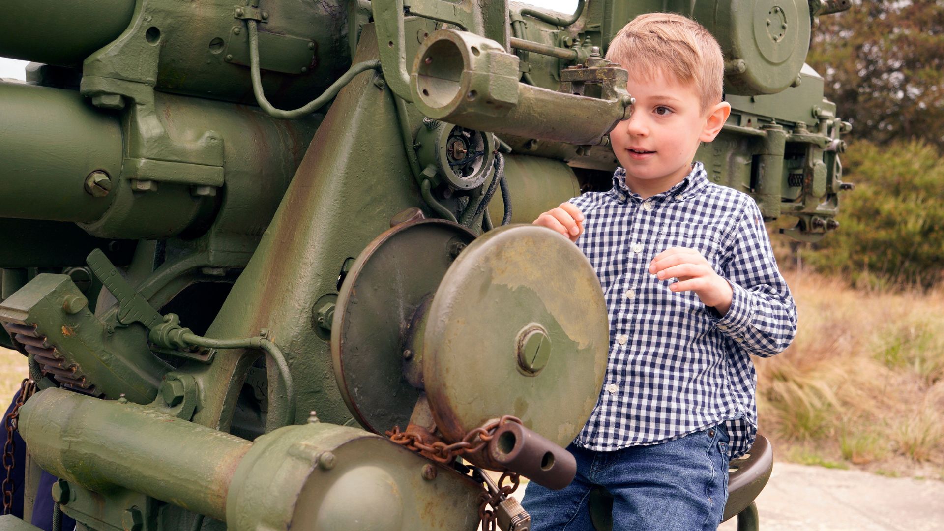 A young boy in a blue-and-white checkered shirt examines the side of a large green military tank outdoors.