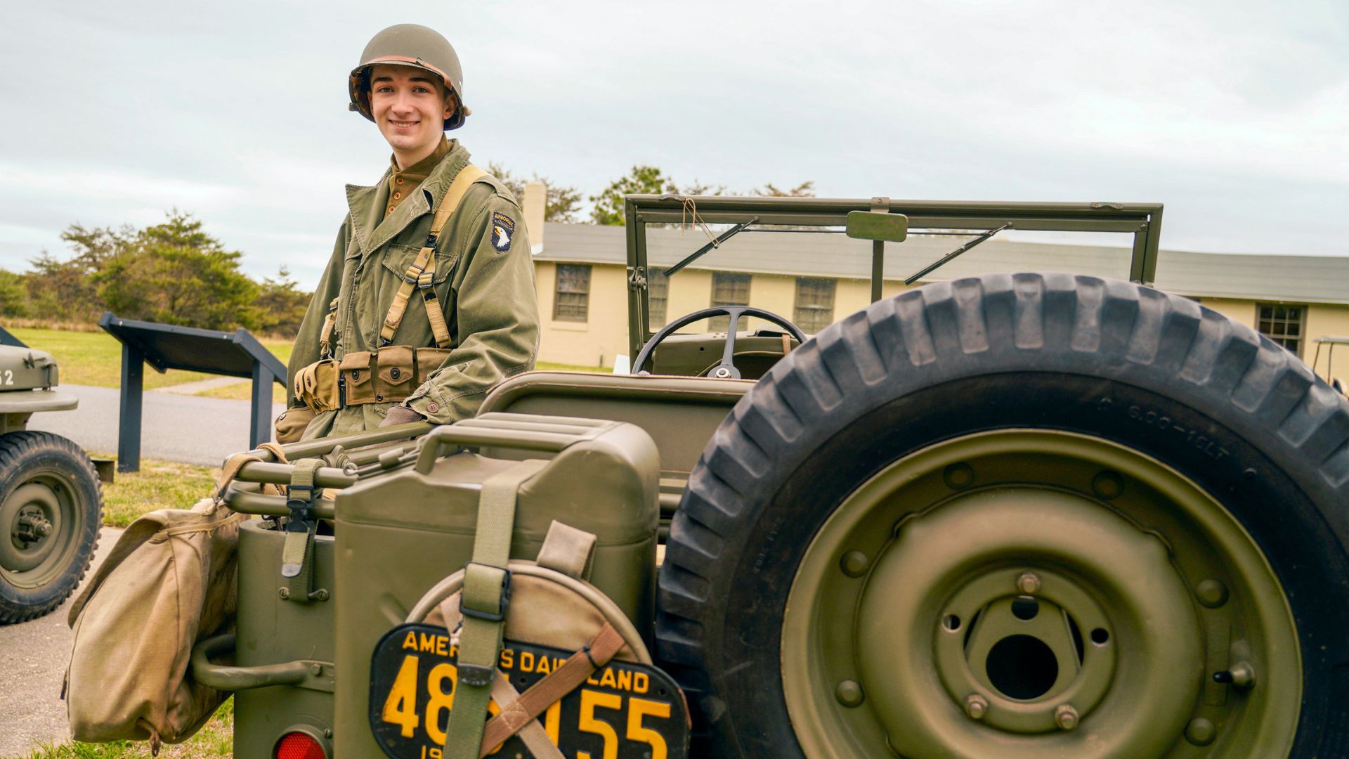 Young man in a WWII-era green uniform wearing a helmet stands beside a vintage olive-drab military jeep, smiling outdoors.