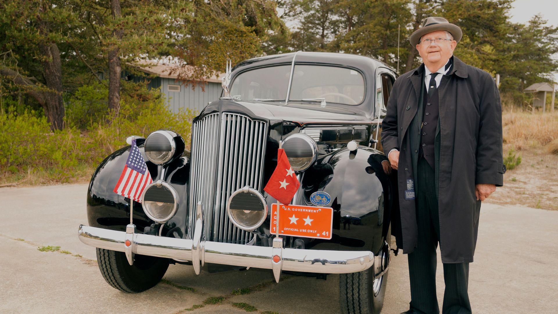 Older man in a suit and fedora stands beside a glossy vintage black car decorated with flags, posing for a photo at a car show.