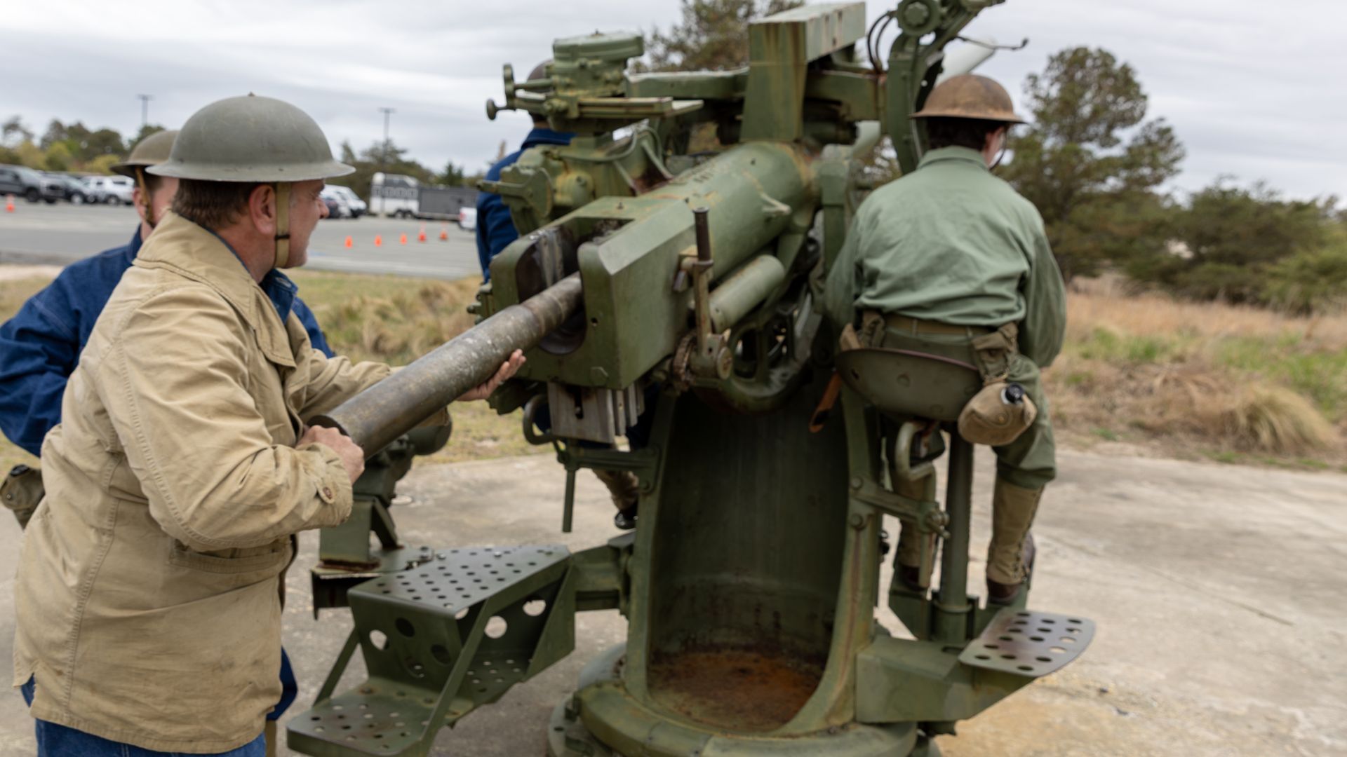 Two soldiers operate a large green field artillery piece outdoors, one loading a shell while another stands nearby.