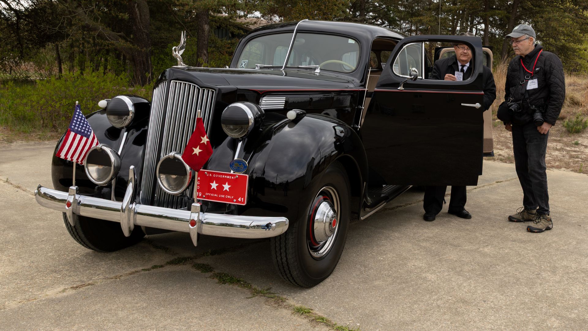 Vintage black luxury car with open passenger door, two men in dark coats beside it, American and red flags mounted on the bumper.