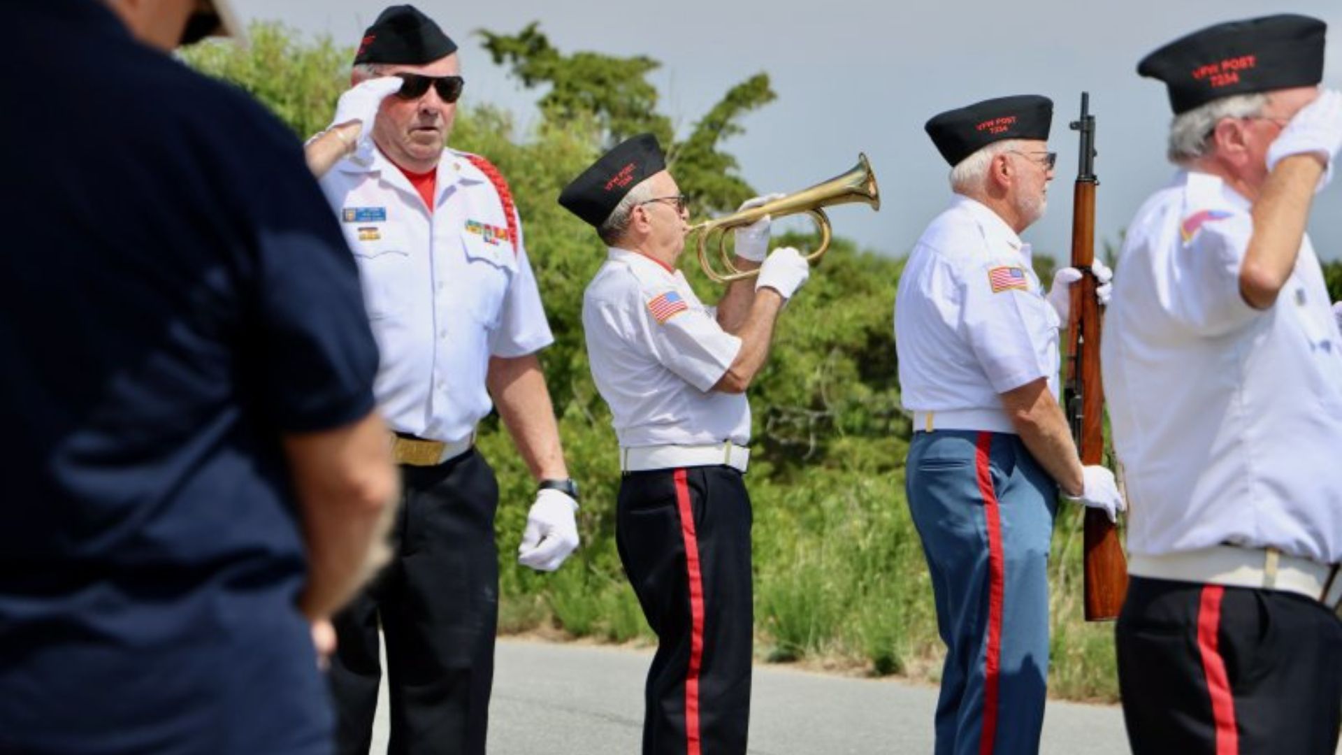 Group of older veterans in white dress uniforms with US flags on sleeves, one man playing a trumpet, others with rifles during a parade.
