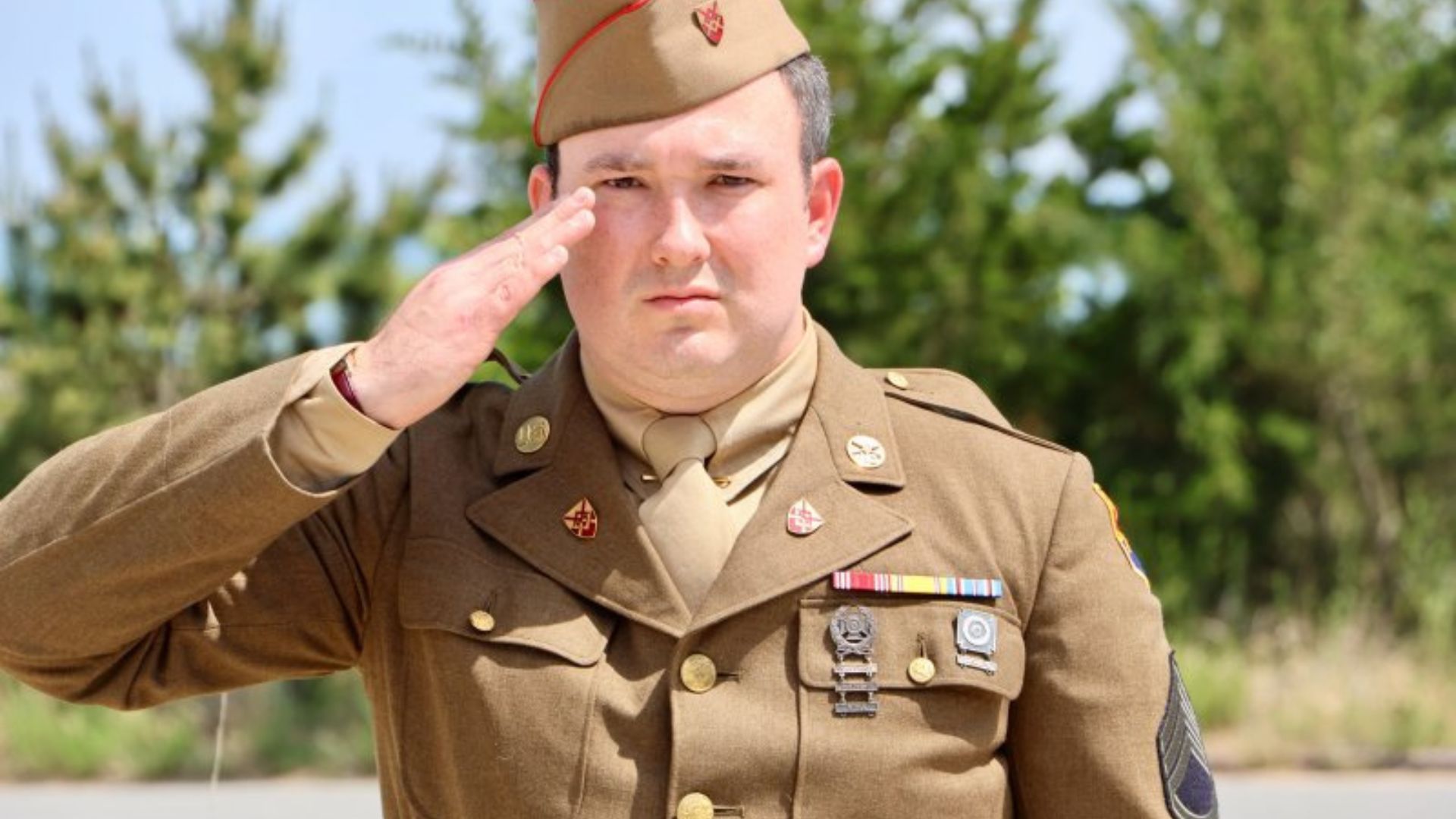 Military officer in tan uniform salutes with his right hand near his temple outdoors.