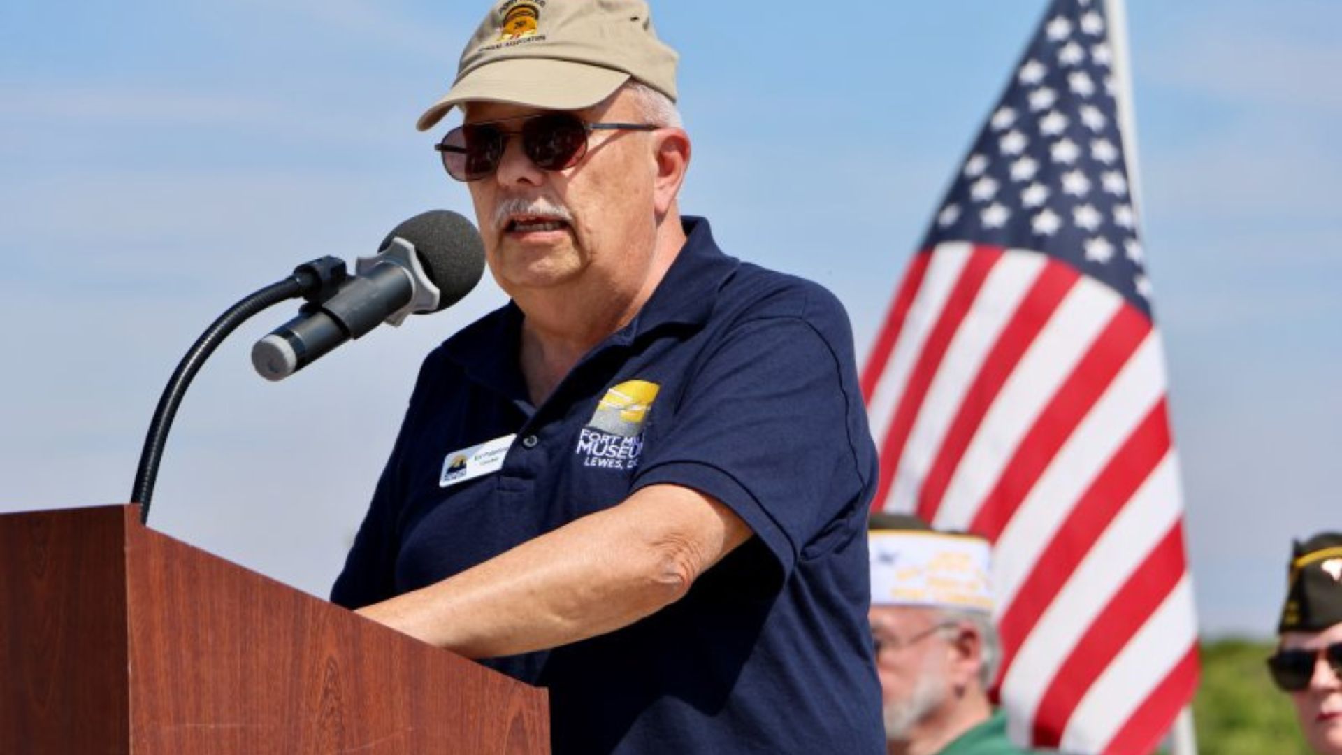 Older man in a beige cap and sunglasses speaks at a wooden podium, microphone in front, with an American flag behind him.