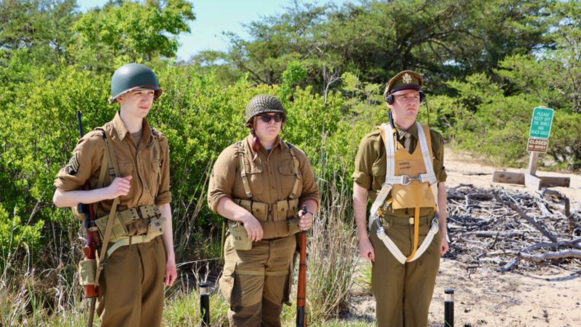 Three people in WWII-era military uniforms pose with gear in a sunny, scrubby outdoor area with driftwood nearby.