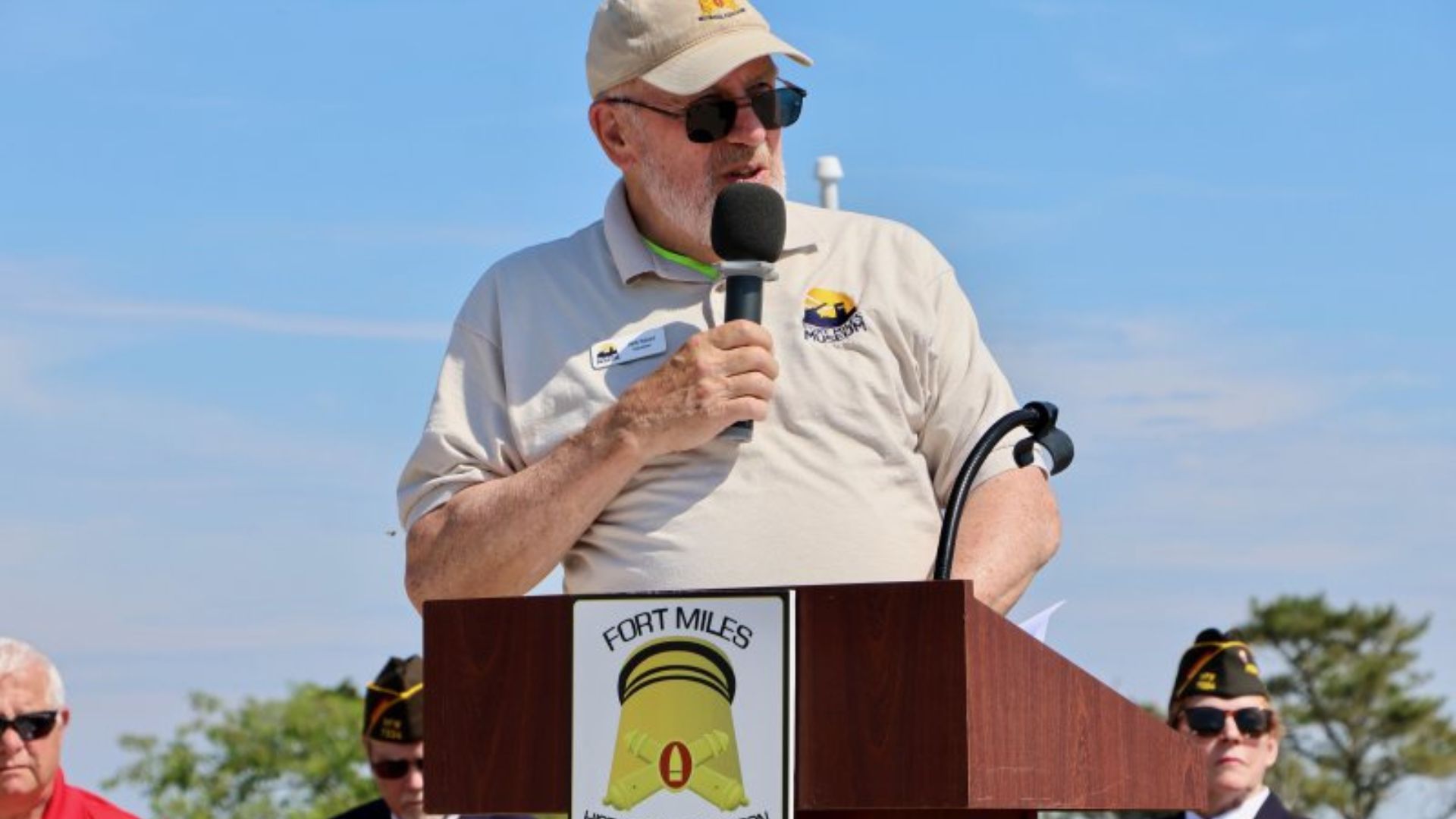 Man wearing a beige polo and cap speaks into a handheld microphone at an outdoor podium, sunglasses on. Proximity shows Fort Miles emblem on the podium badge.