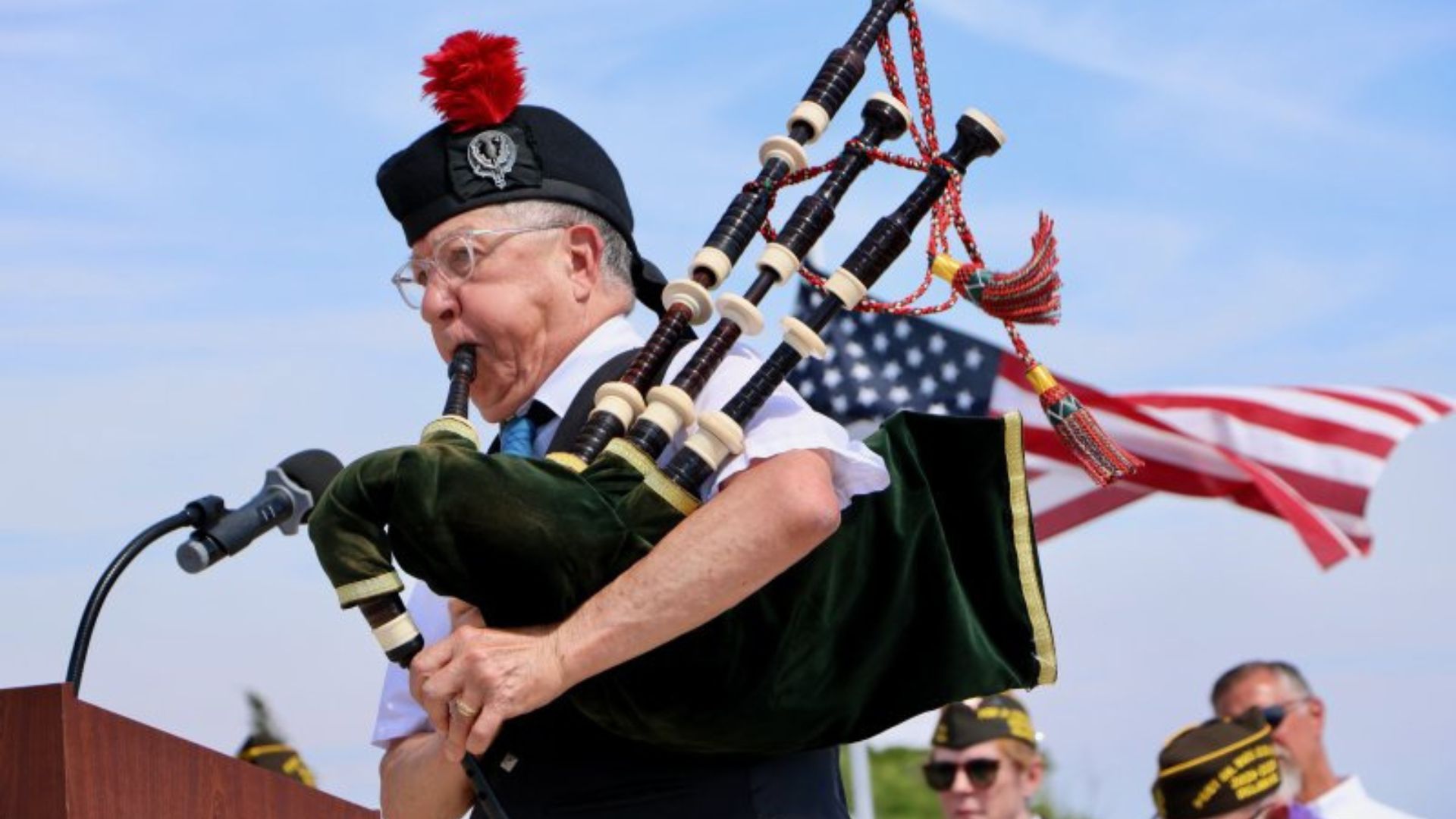 Man playing bagpipes at an outdoor ceremony, wearing a black hat with a red plume, beside a wooden podium and an American flag in the background.