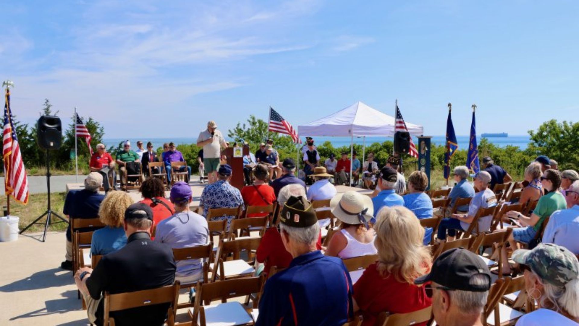 Outdoor ceremony on a sunny day by the coast with an audience seated in rows and a speaker at a podium under a white canopy; American flags line the area.