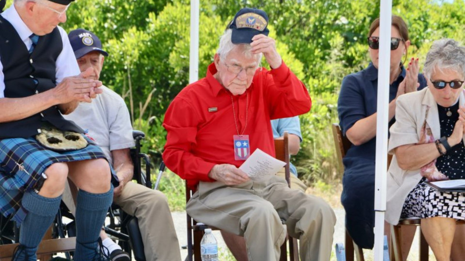 Elderly veterans sit outdoors; man in a red shirt reads a paper while adjusting his cap as others applaud.