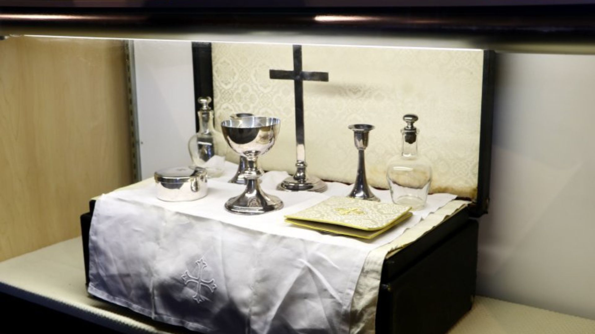 Silver chalice, cruets, and liturgical vessels arranged on a white cloth altar with a cross backdrop inside a display cabinet.