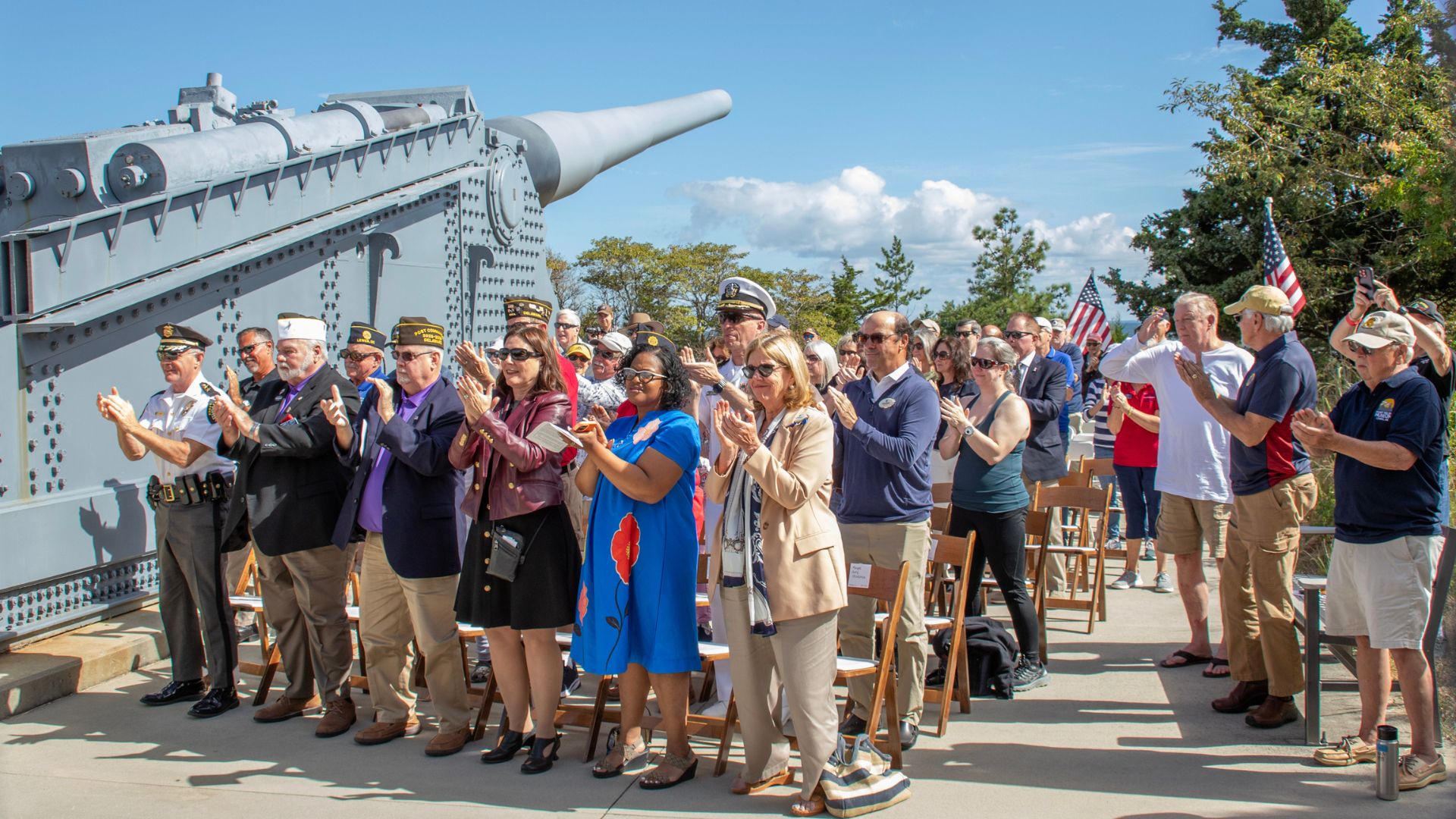 Group of veterans and civilians applauding at a ceremony beside a large naval gun on a bridge, with American flags in the background on a sunny day.