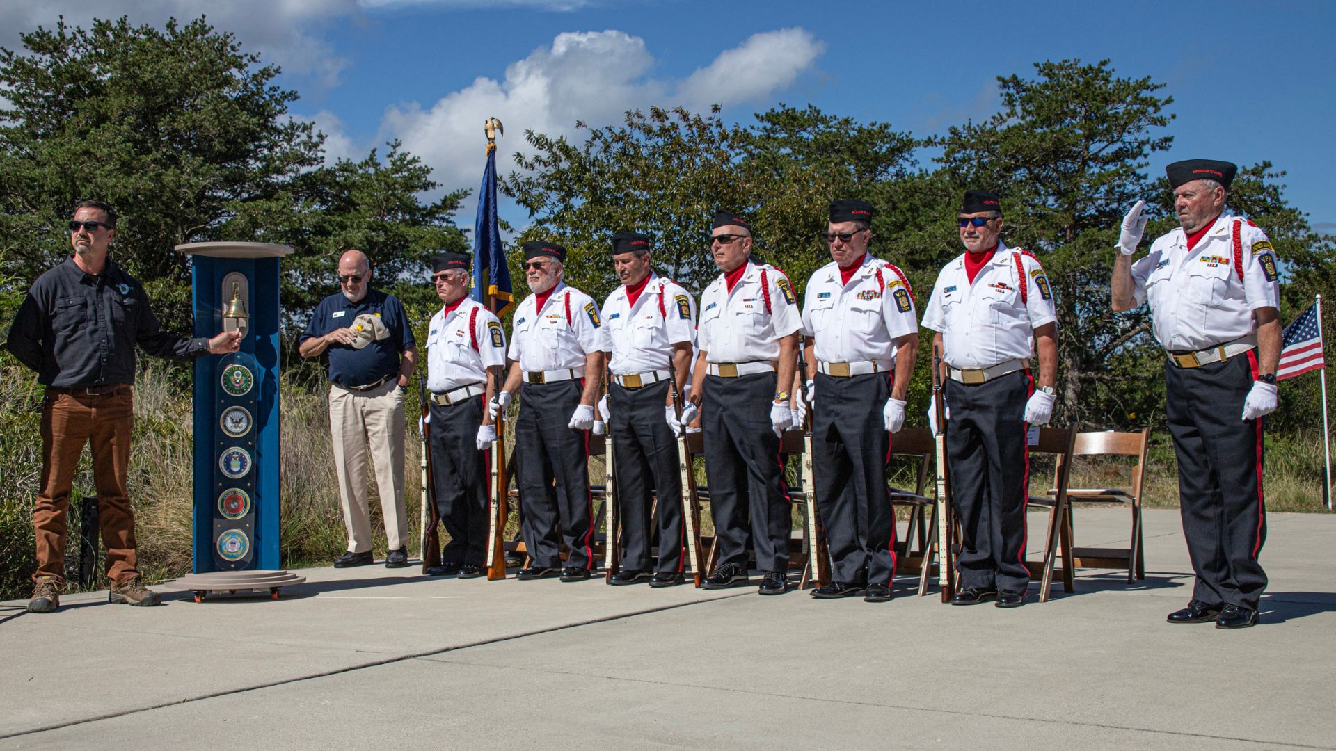 Veterans in white dress uniforms standing in a line for a ceremony, with one man ringing a bell on a blue column to the left.