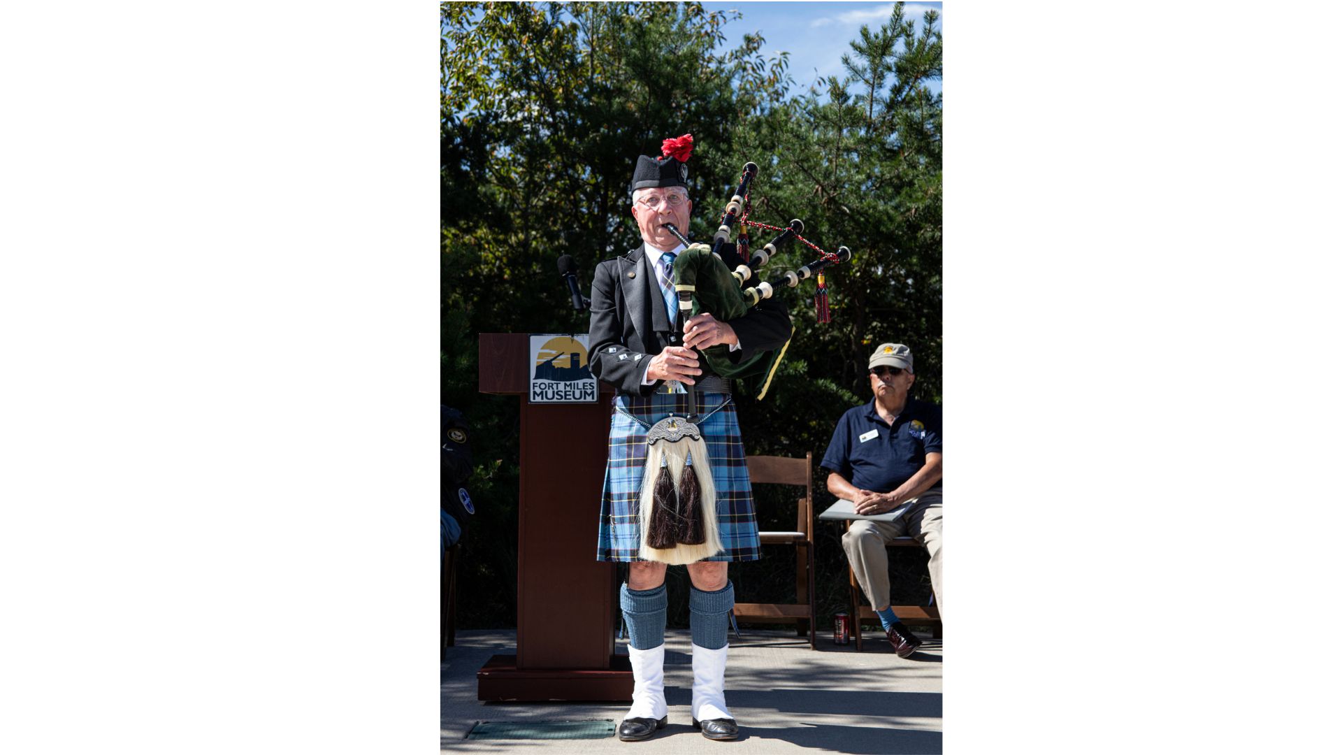 Man in a blazer and kilt playing bagpipes outdoors, with a Fort Miles Museum sign in the background.