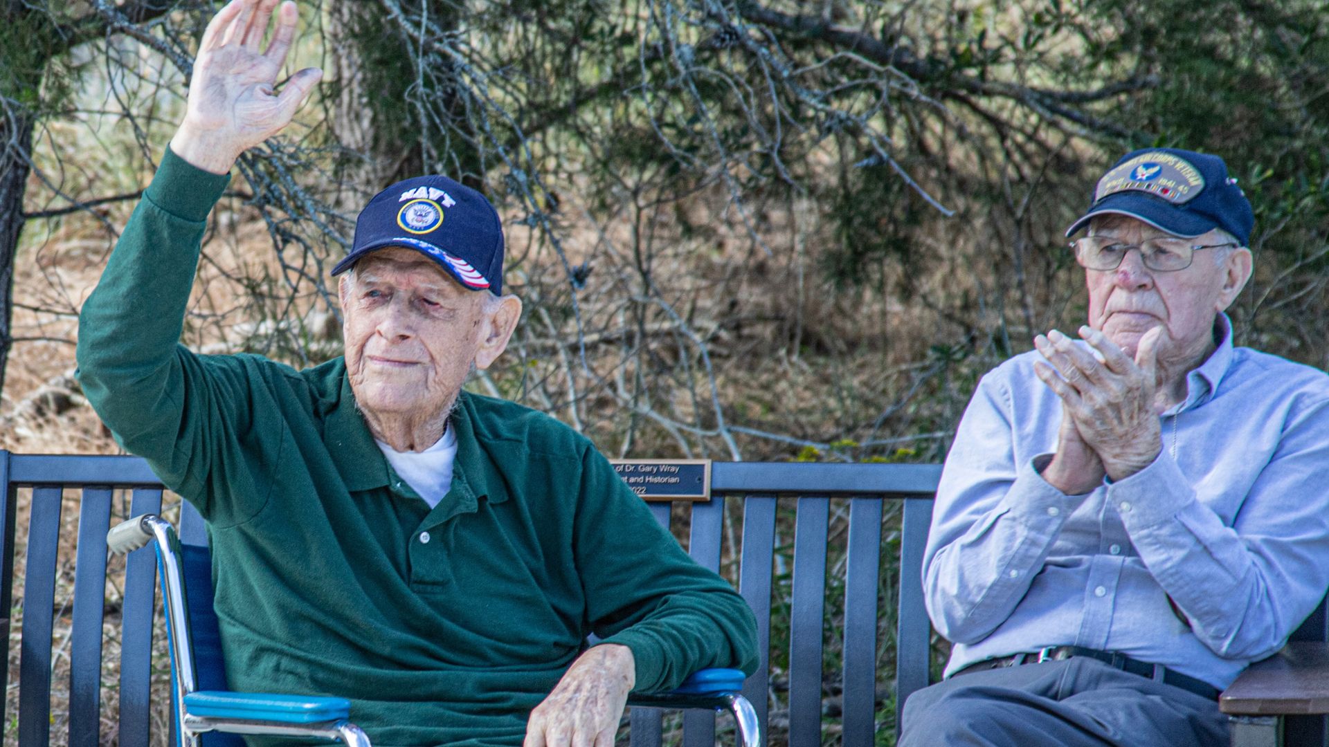 Two elderly men sit on a park bench outdoors; one raises his hand to wave while the other clasps his hands together.