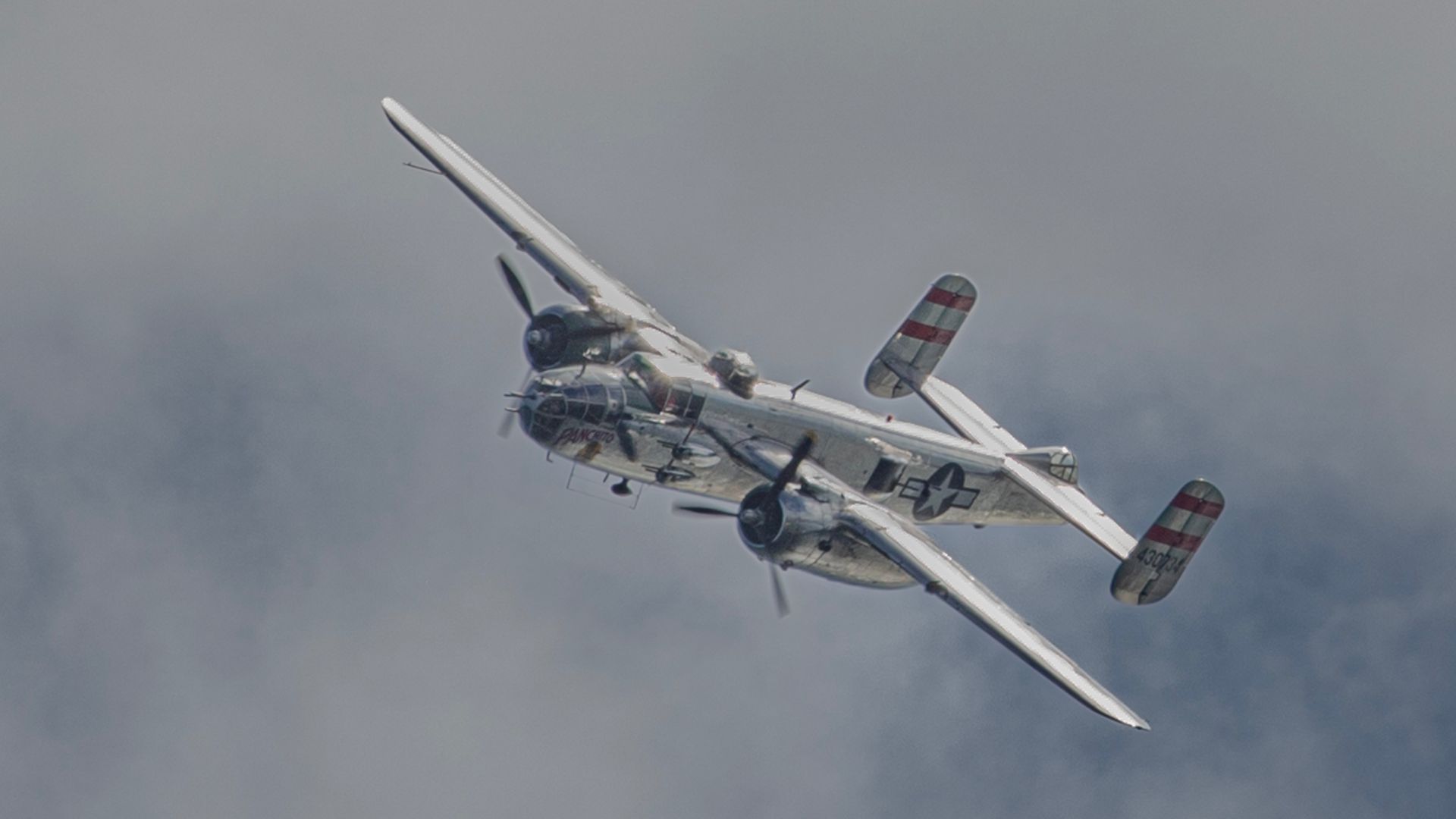 Silver WWII-era twin-engine bomber in mid-flight against a cloudy sky with rotating propellers visible.